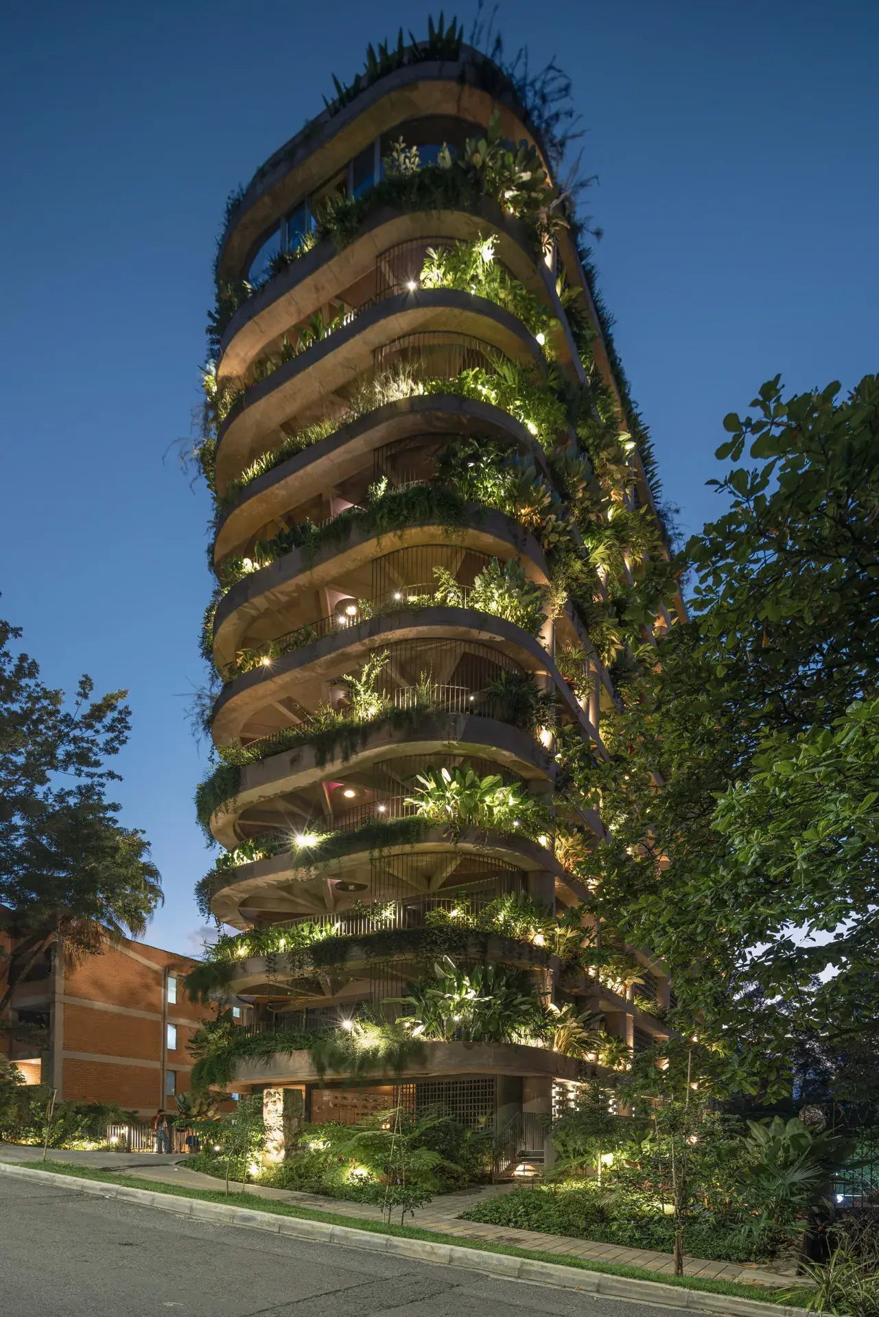 Torre residencial 23 Living de PlanB Arquitectos al atardecer, con balcones curvos cubiertos de vegetación e iluminación cálida en Medellín.